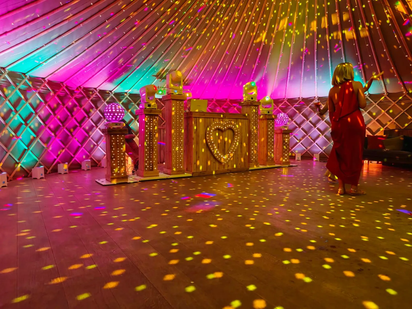 Yurt dance floor lit in pink and gold with a guest dancing beside the rustic DJ booth